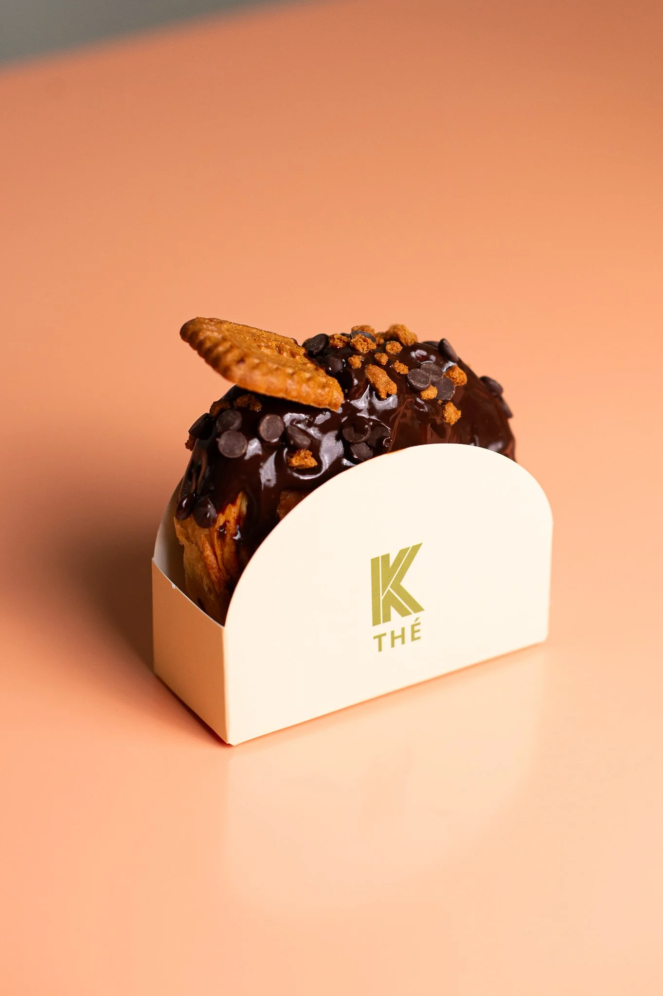 Un donut au chocolat glacé avec des pépites de chocolat, une crème ou sauce au centre, surmonté d'un biscuit et d'une petite miette, présenté dans un support blanc avec un logo vert 'K THÉ' sur fond beige.