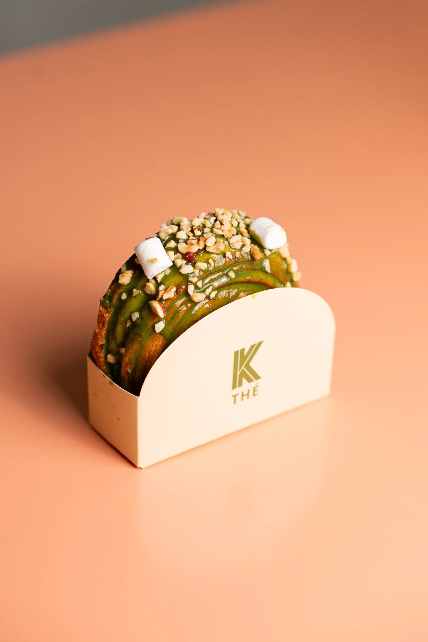 Une pâtisserie de forme rectangulaire avec une garniture verte, décorée de noisettes hachées, de marshmallows blancs, et posée dans un support blanc portant le logo 'K THÉ' sur un fond rose.