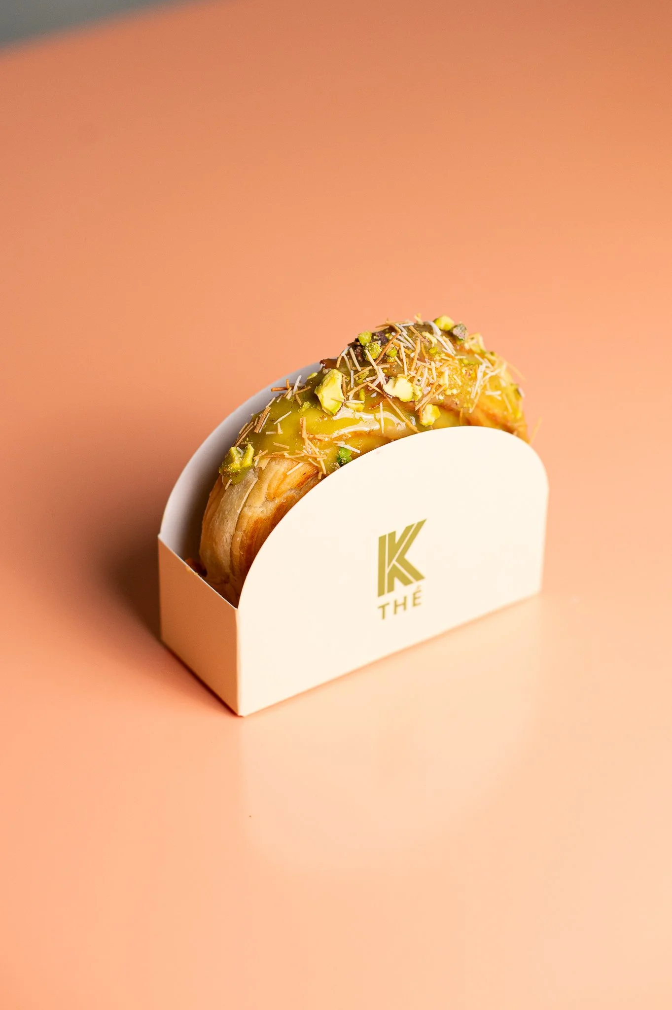 Un croissant garni de crème, pistaches, éclats de chocolat, dans une boîte blanche avec un logo 'K THÉ', placé sur une surface rose