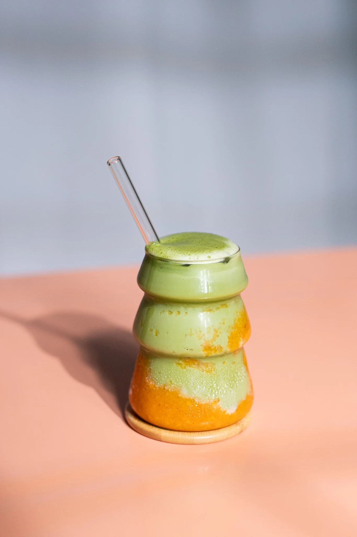 Verre en céramique en forme de poupée chinoise, rempli d'une boisson vert vif, avec une paille en verre, posé sur une surface rose.
