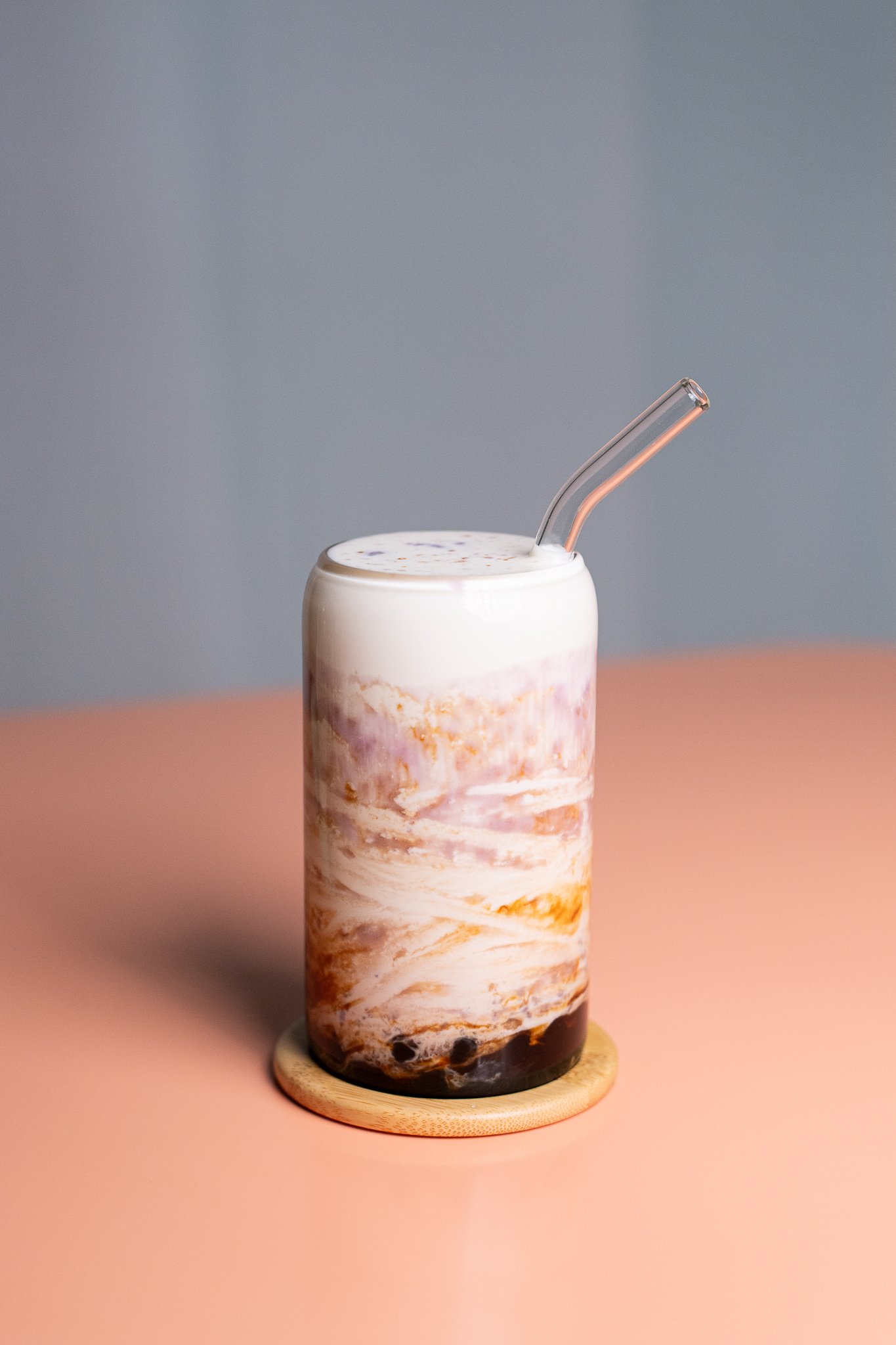 Un verre de boisson, probablement un milkshake ou un thé glacé, avec un couvercle blanc, un mélange de couleurs brunes, roses et blanches, et une paille en plastique clair, posé sur un dessous en bois sur une surface rose pâle, avec un arrière-plan gris.