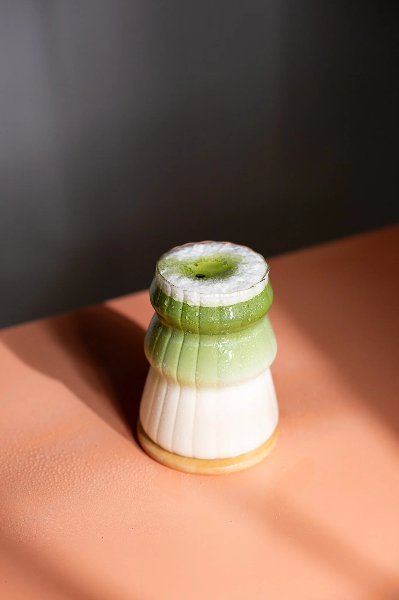 Une bouteille de savon ou contenir à la vie en forme de lézard avec des coloris dégradés de blanc, vert et beige, placée sur une surface rose au fond noir.