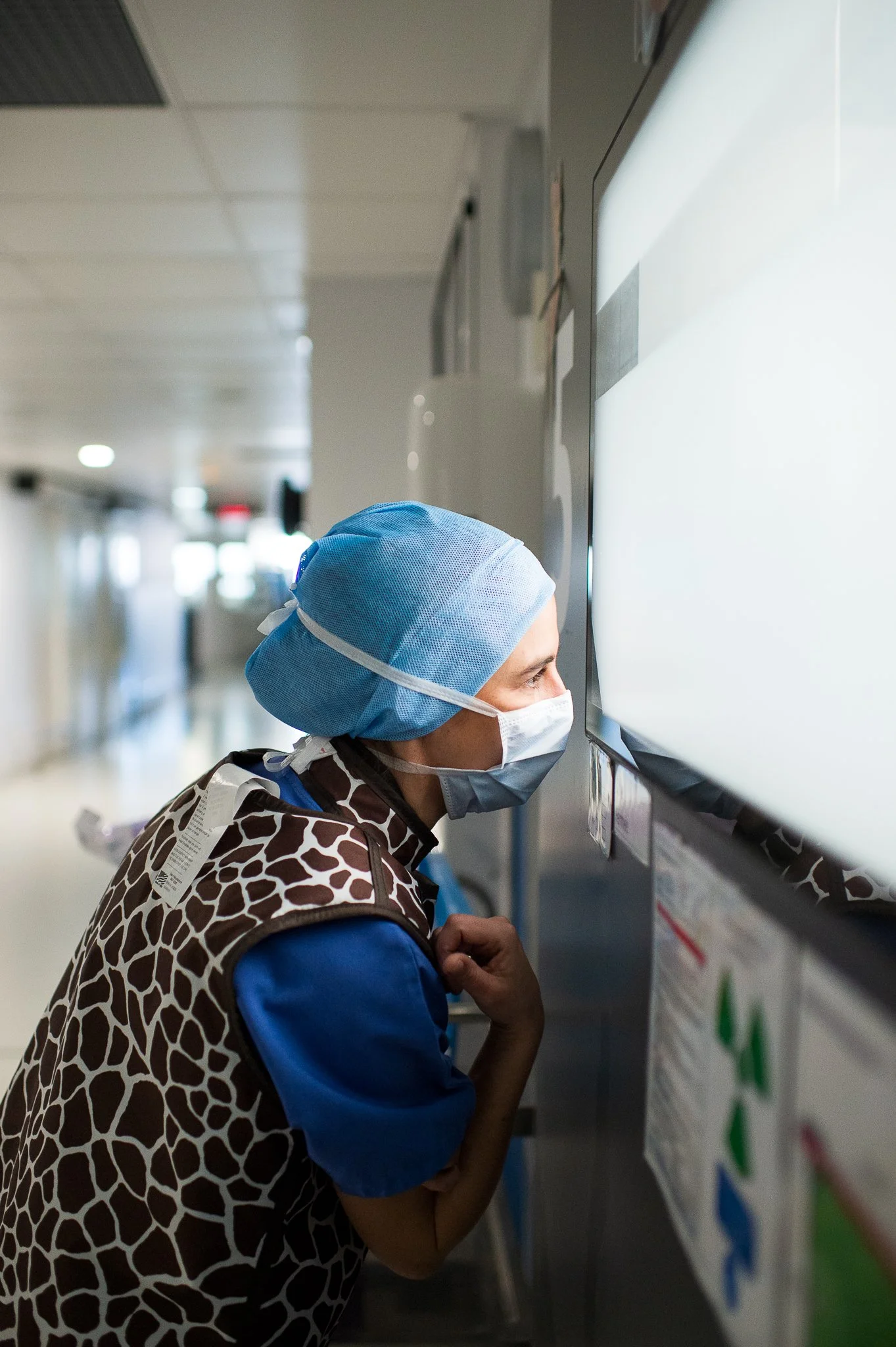 Un personnel médical portant un masque, une coiffe et un gilet avec un imprimé girafe, regarde un écran dans un couloir d'hôpital.