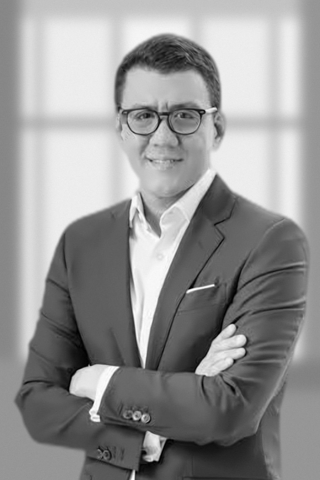 Black and white professional portrait of a man with glasses, wearing a suit and white shirt, smiling with arms crossed.
