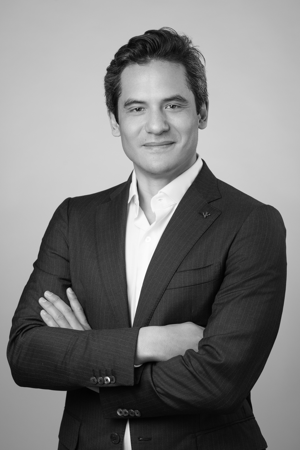 A black and white headshot of a man in a business suit with arms crossed, smiling slightly, against a plain background.