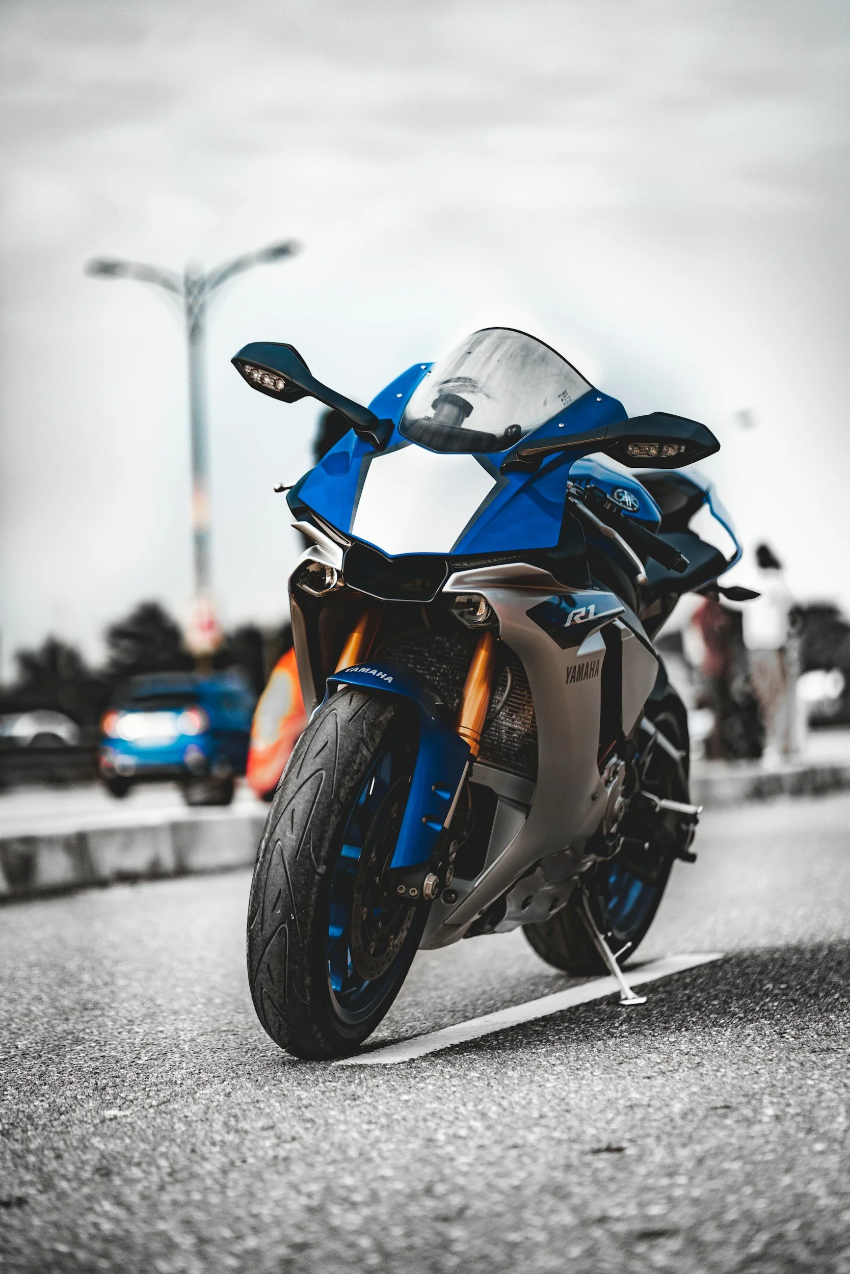 A blue and silver Yamaha sport motorcycle parked in a parking lot during daytime, with blurred vehicles and lamp posts in the background.