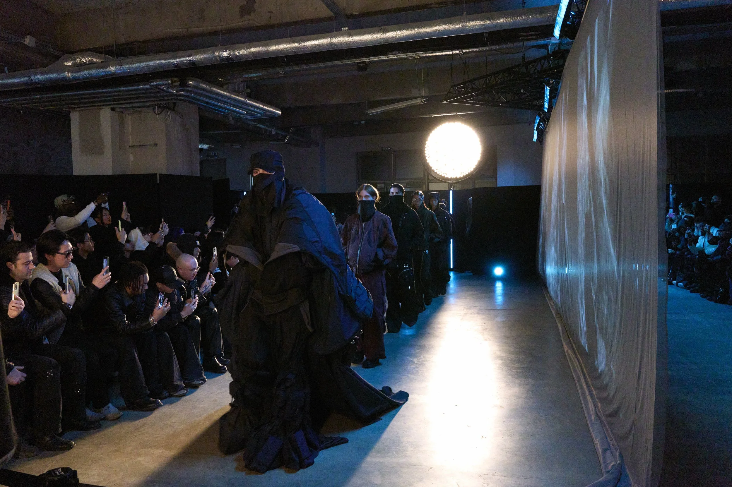 Défilé de mode sombre avec mannequins masqués, public photographiant et éclairage bleu dans un espace industriel.