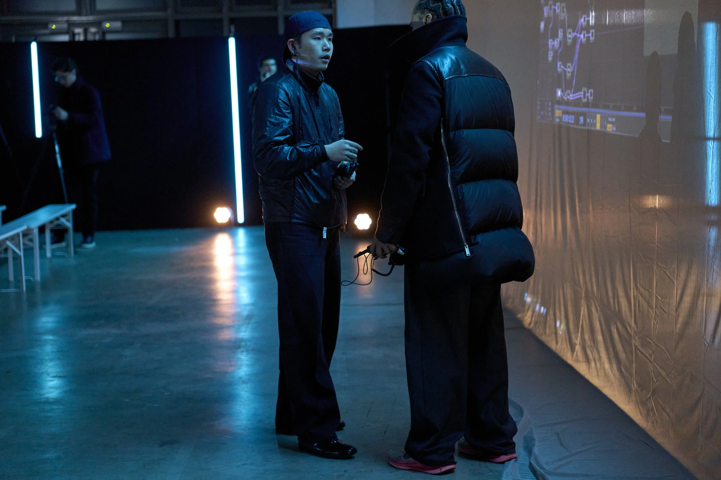 Deux personnes en vêtements noirs discutent dans un espace sombre éclairé par des néons bleus et des projections numériques.