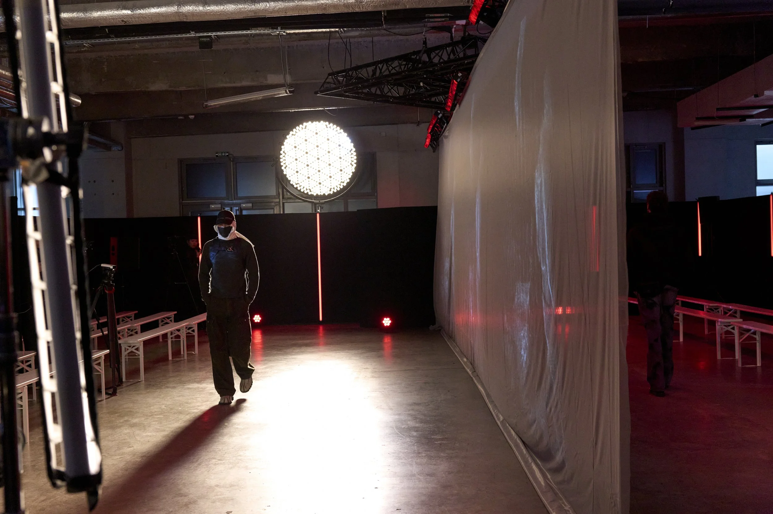 Coulisses d’un événement avec éclairage scénique rouge, projecteurs, gradins et technicien portant un masque.