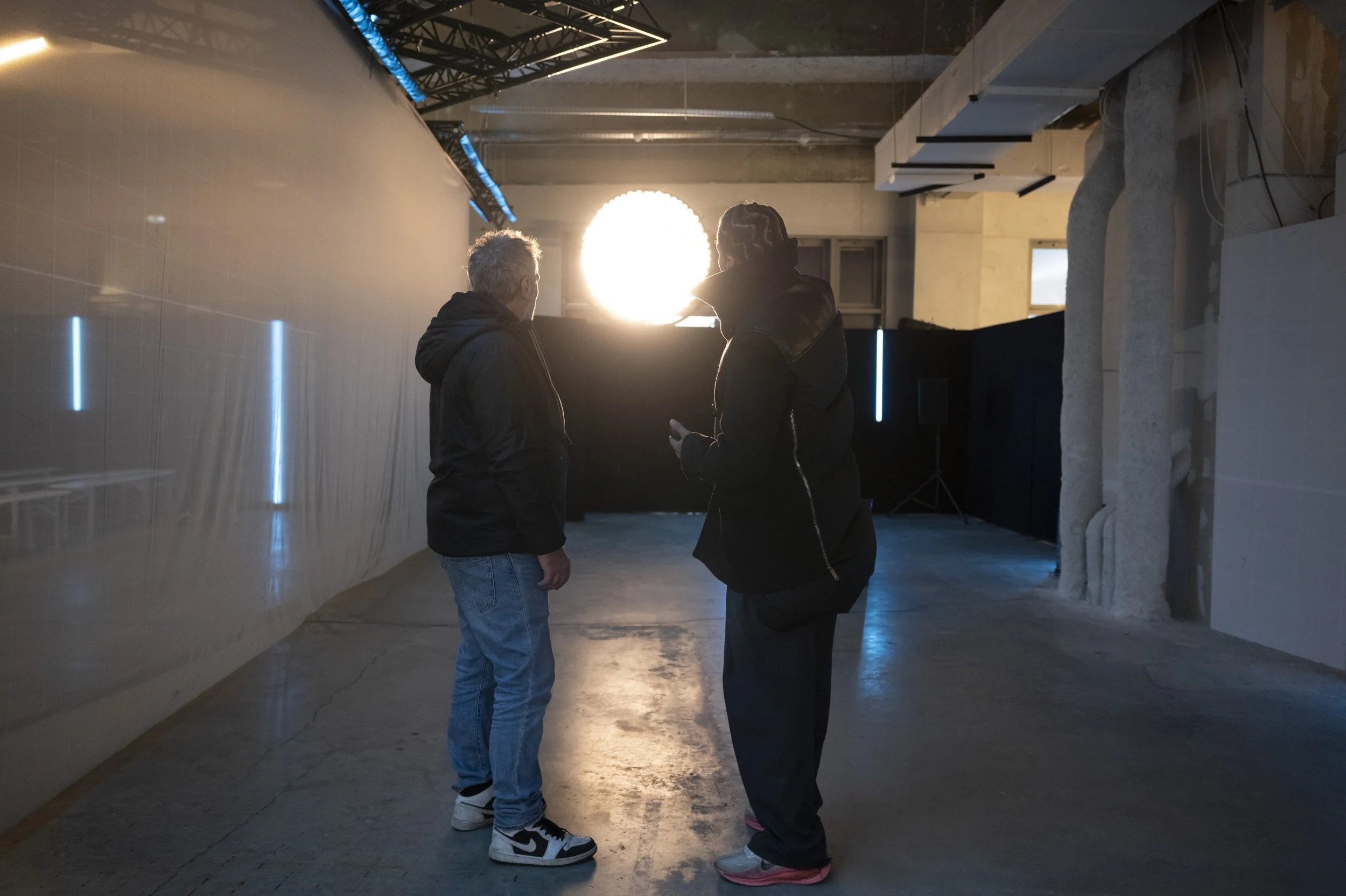 Deux personnes discutent dans un espace industriel éclairé par des néons bleus et un projecteur lumineux.