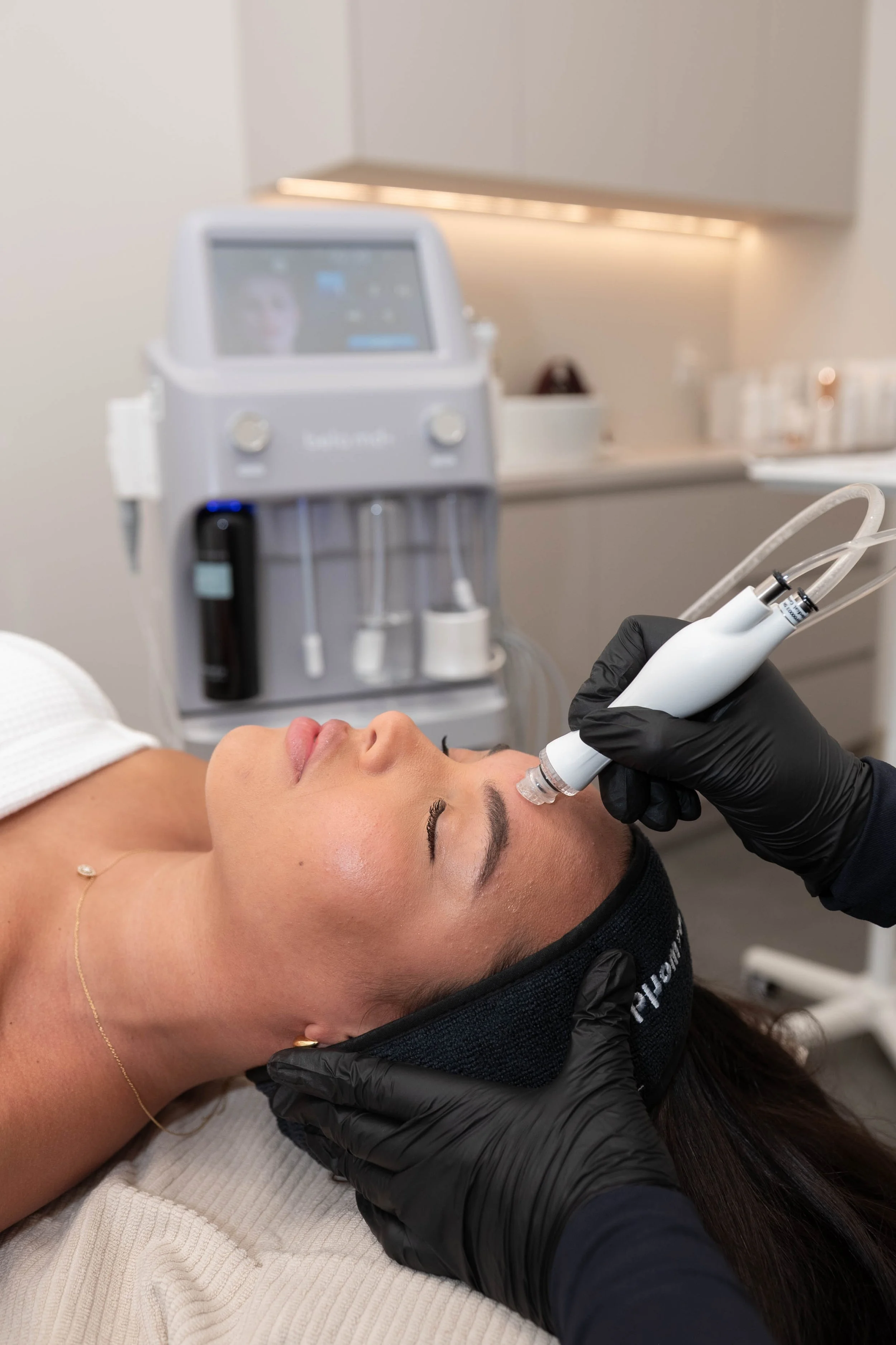 Une femme reçoit un traitement esthétique du visage avec un appareil médical, allongée sur une table, dans une clinique ou un salon de beauté. Une technicienne porte des gants noirs et tien un dispositif qui applique un traitement sur le visage de la patiente.