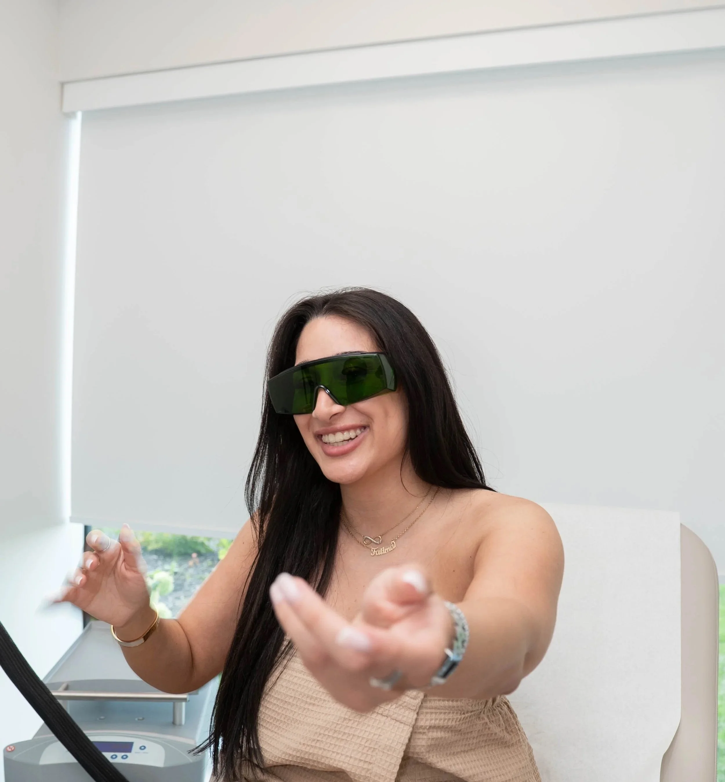 Une femme souriante portant des lunettes de soleil de réalité virtuelle, vêtue d'une robe beige sans manches et portant des bijoux, pose devant un miroir dans une pièce lumineuse avec une grande fenêtre en arrière-plan.