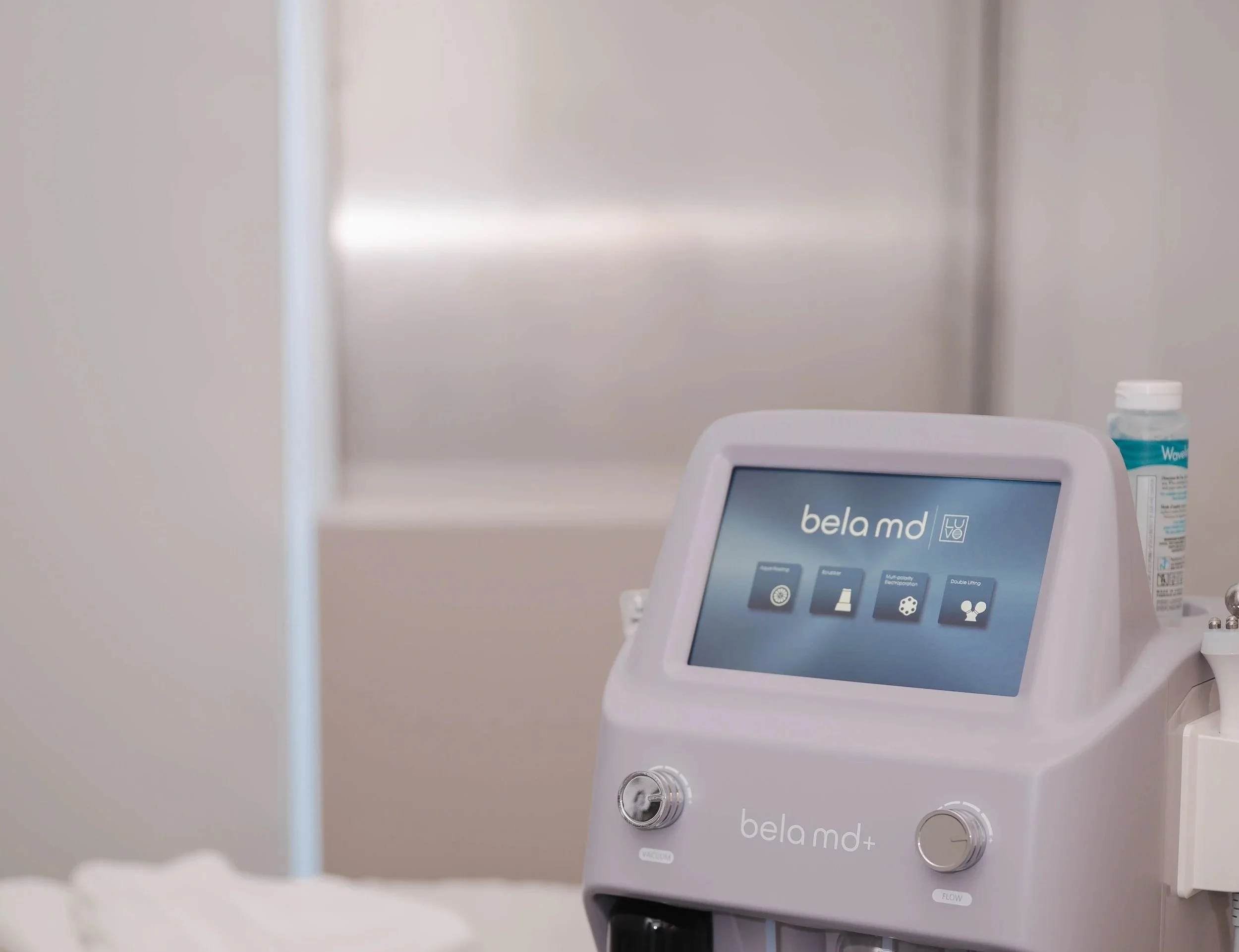 Un appareil médical nommé Bela MD+ avec un écran tactile affichant un menu. Appareil blanc avec des boutons et des câbles connectés, placé sur une surface dans une pièce blanche ou un environnement médical.
