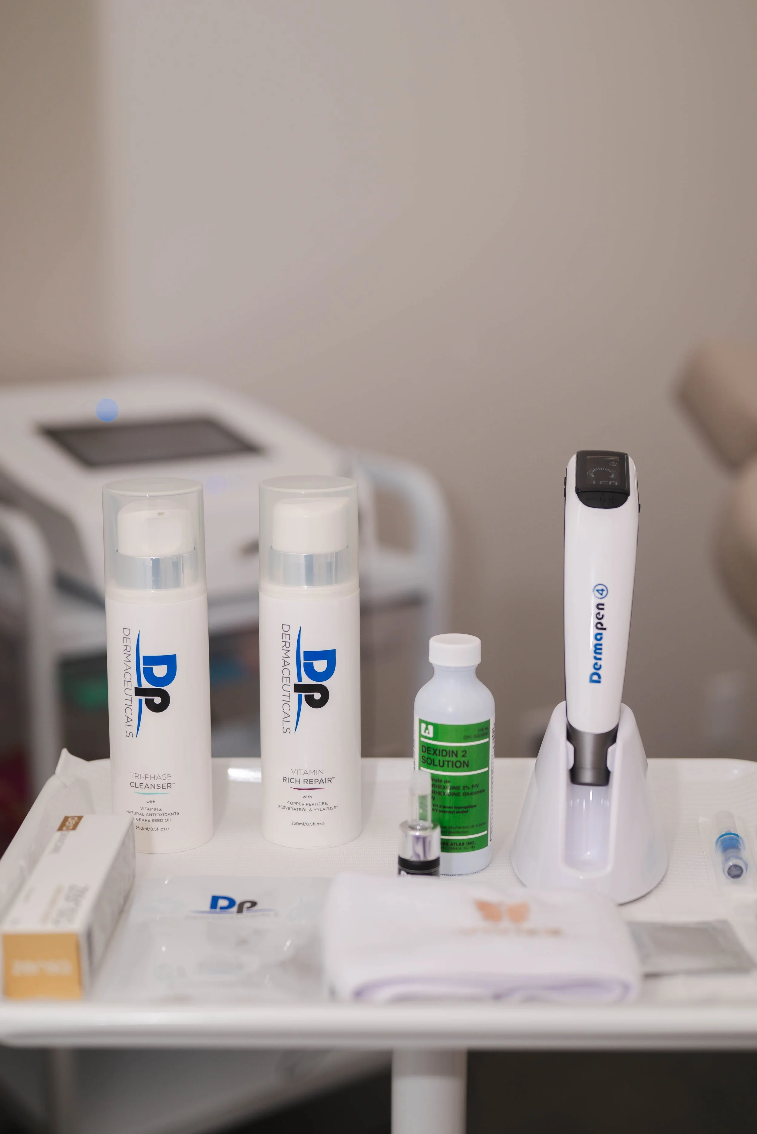 Produits cosmétiques et médicaux sur un plateau de table, notamment deux tubes de crème de dermo-cosmétique, un flacon de solution curet, une brosse électrique, un gel désinfectant, et un emballage de coton dans un environnement de clinique ou de laboratoire.
