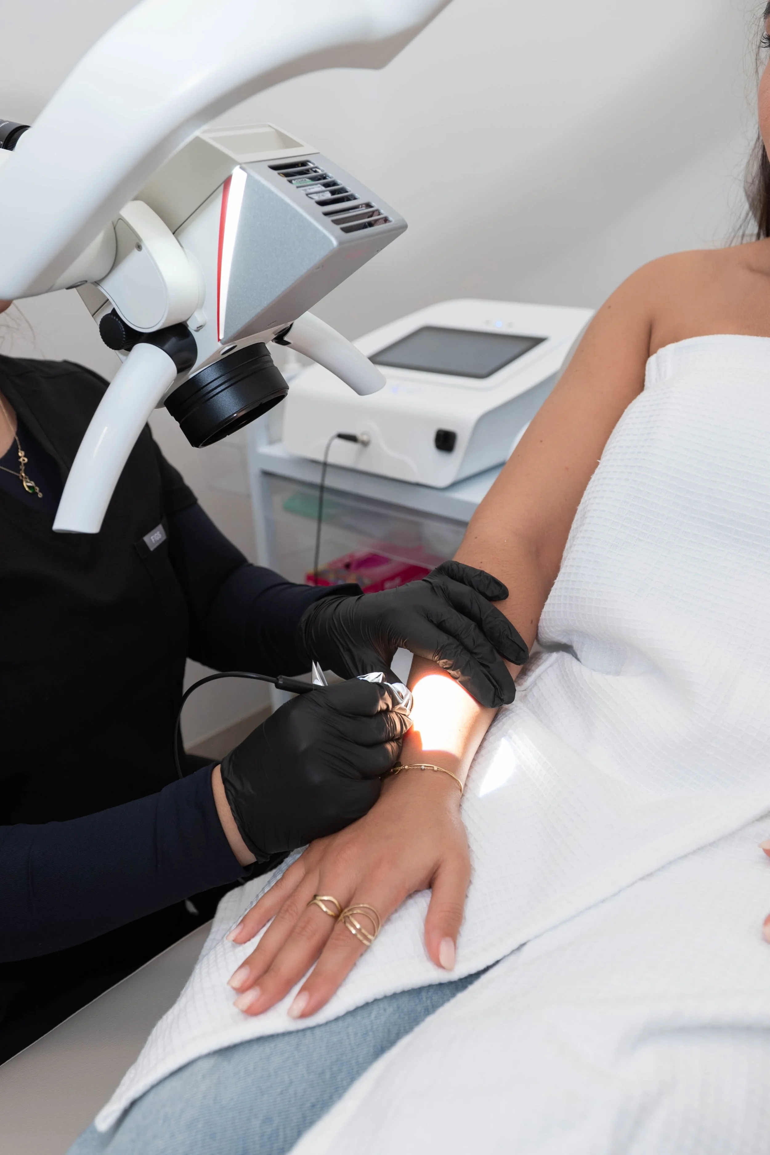 Une femme reçoit une injection dans le bras par une professionnelle de santé. La femme est allongée sur une table, portant une robe blanche et des bijoux, tandis que la professionnelle porte des gants noirs et utilise un appareil médical pour la procédure.