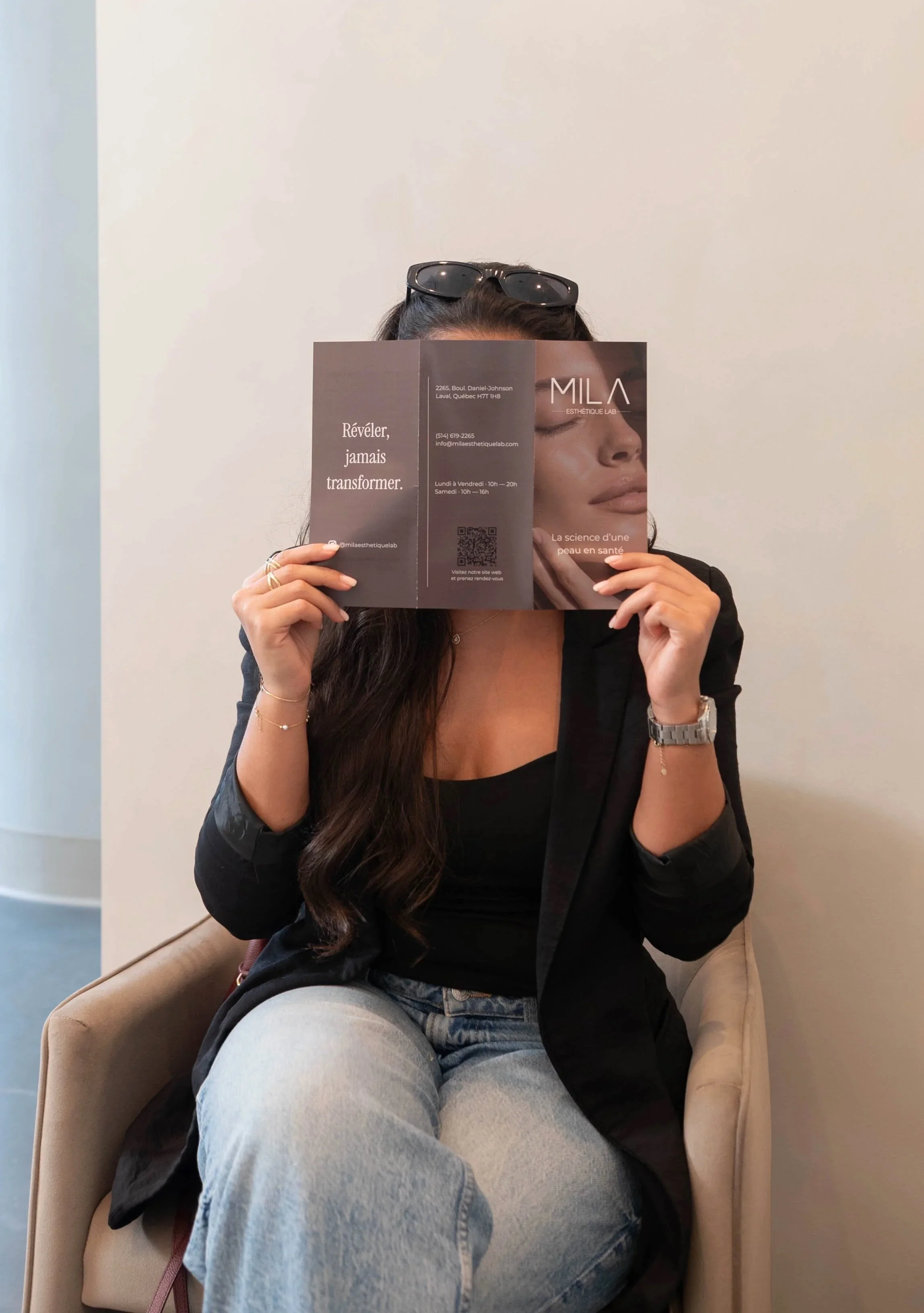 Une femme assise dans un fauteuil beige, portant un jean gris et un haut noir, tient un magazine devant son visage, avec des lunettes de soleil sur sa tête. Elle porte des bracelets et une montre.