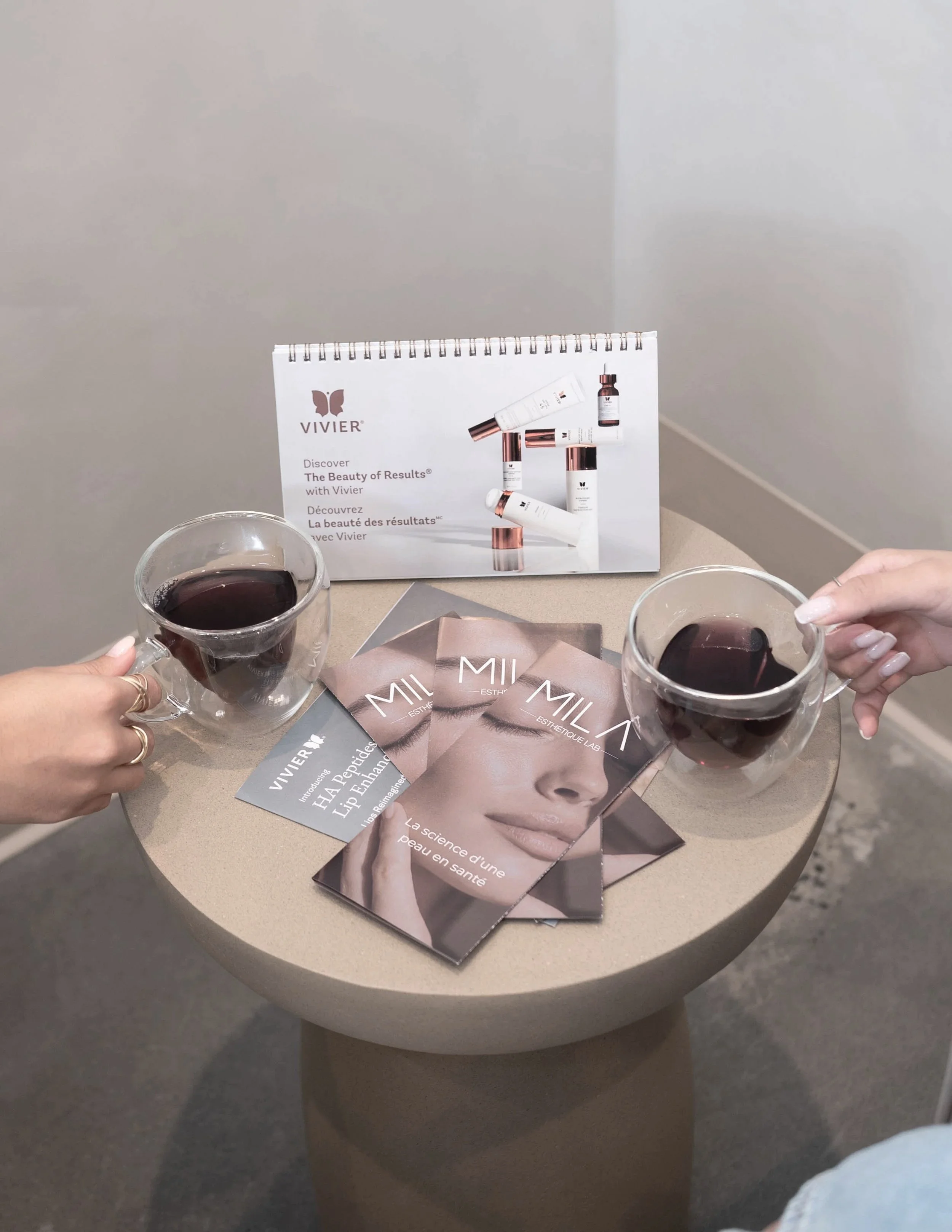Deux mains tenant des verres de vin rouge au-dessus d'une table ronde avec des brochures promotionnelles d'une clinique esthétique. En arrière-plan, un calendrier avec le logo VIVIER et une image de visage féminin, ainsi que des brochures intitulées 'MIL' et 'Esthétique Lab'.