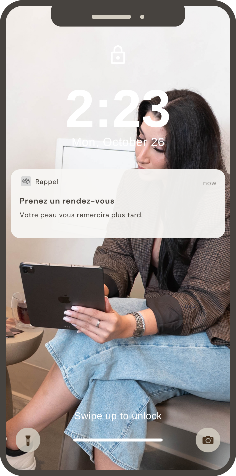 Capture d'écran d'un téléphone affichant une femme utilisant une tablette, avec une notification rappel pour prendre rendez-vous, portant une chemise à carreaux et un jean, assise dans un espace intérieur.