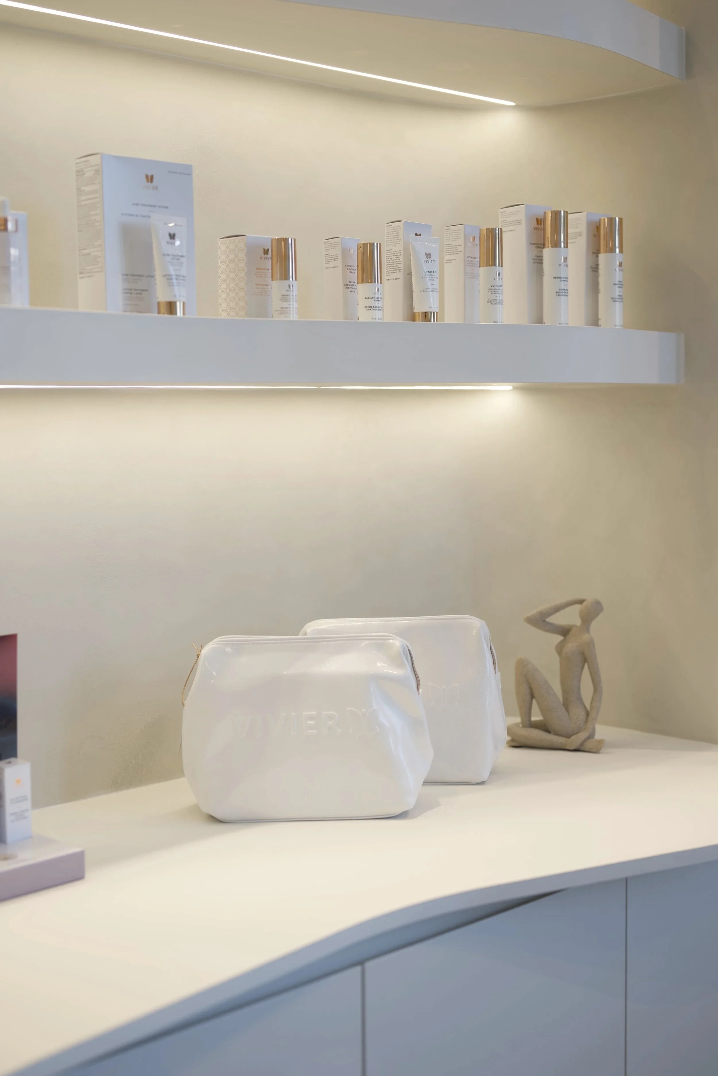 Display de produits de soins sur une étagère dans un décor moderne, avec deux sacs en plastique blancs sur le comptoir et une sculpture abstraite en bois ou pierre à côté.
