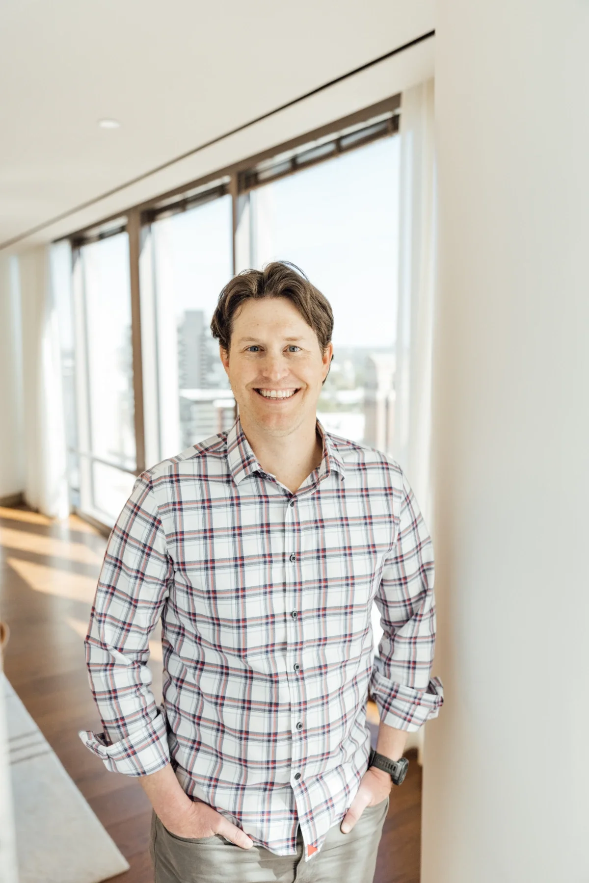 Smiling young man with brown hair wearing a plaid shirt standing indoors near windows and a white wall.