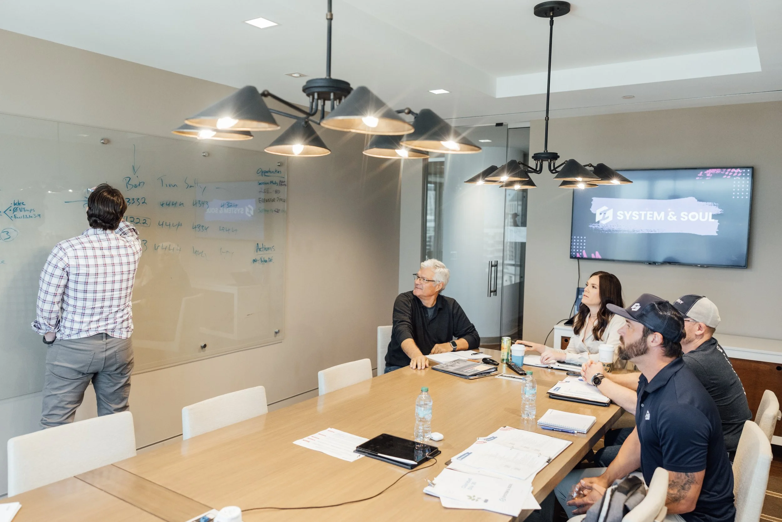 A group of people attending a meeting in a conference room. One person is standing at a whiteboard writing notes, while others are seated around a large table listening. There is a large screen on the wall displaying 'System & Soul' and some documents and water bottles on the table.