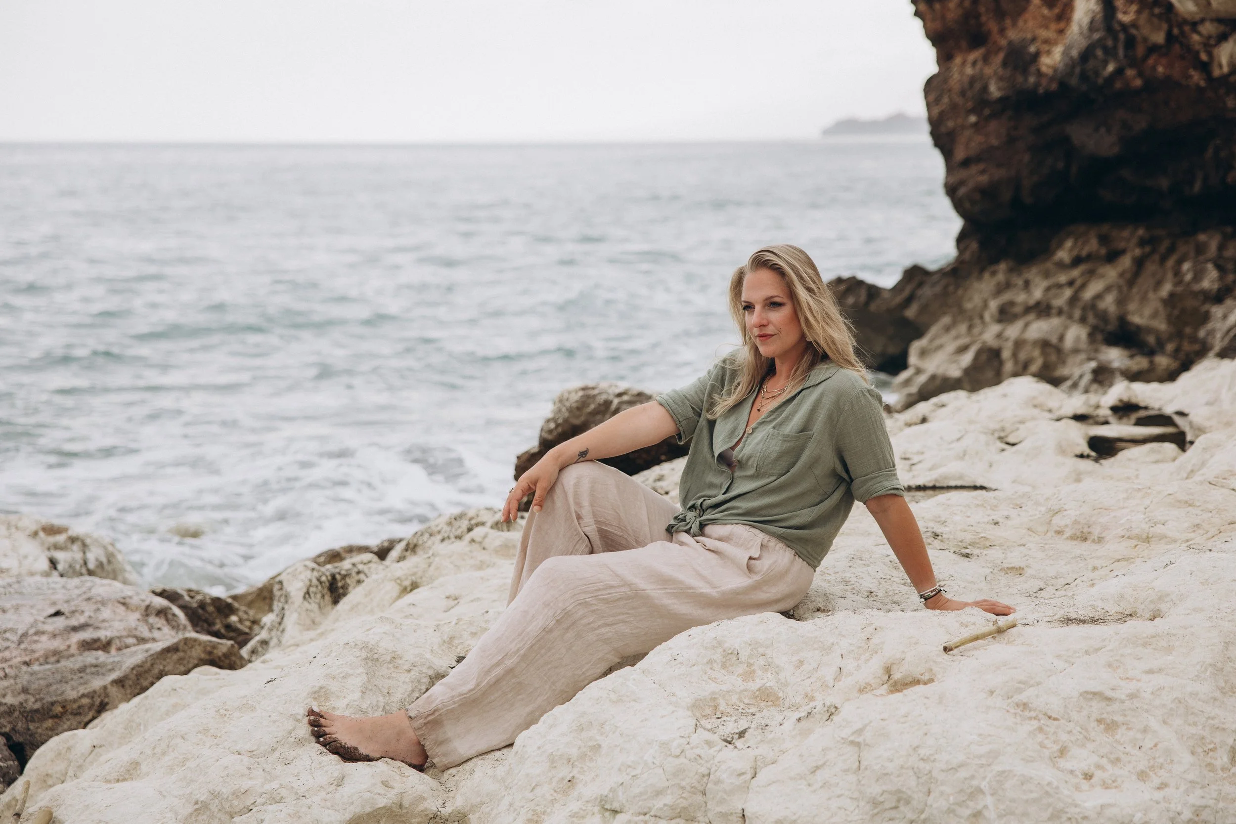 Een vrouw zit op rotsen aan de kust, met de oceaan en een rotsformatie op de achtergrond.