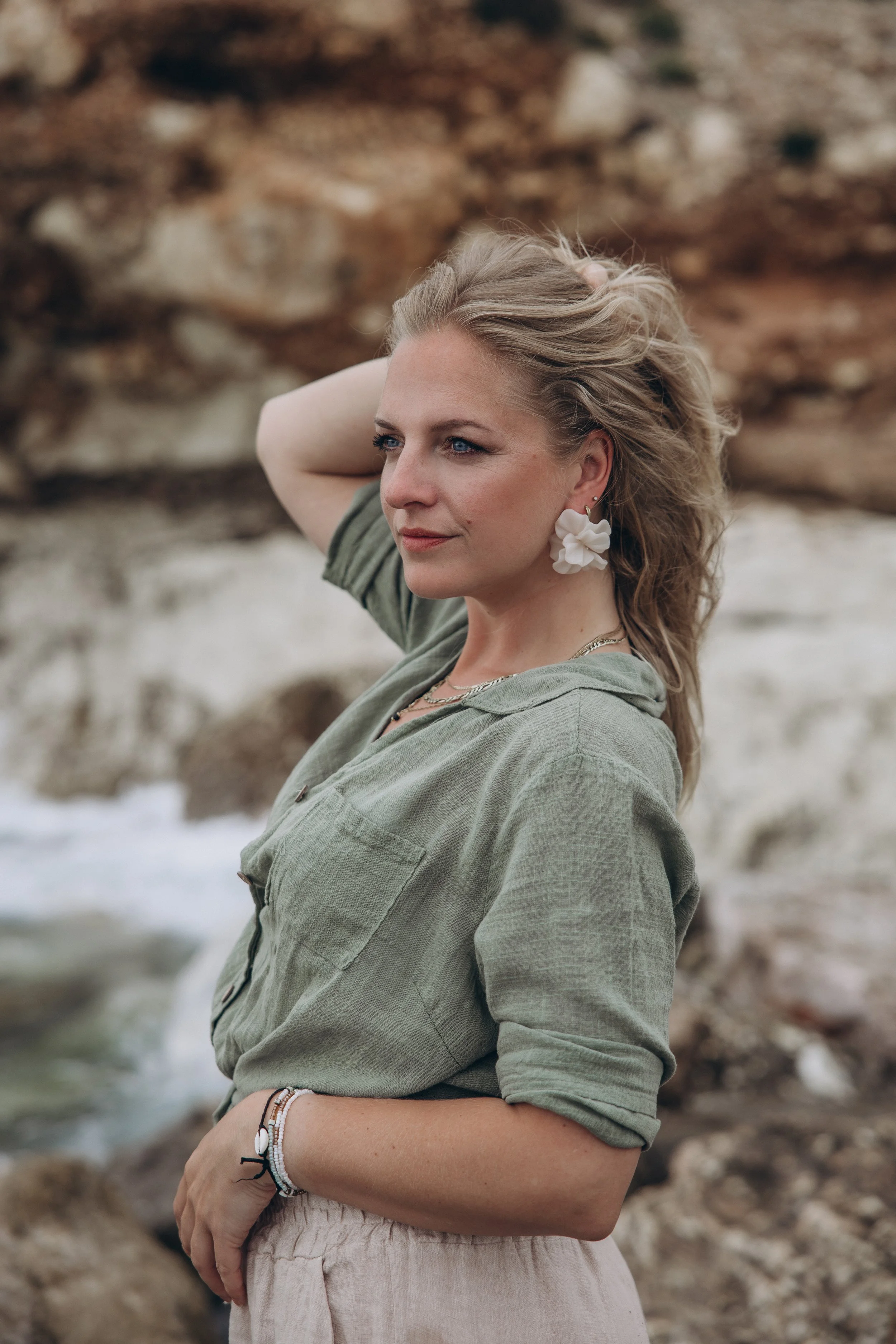 Een vrouw met lange, golvende blonde haar die rust op haar schouders, draagt grote witte bloemoorbellen, een groene blouse en armbanden, en staat aan een strand met rotsen en water op de achtergrond.