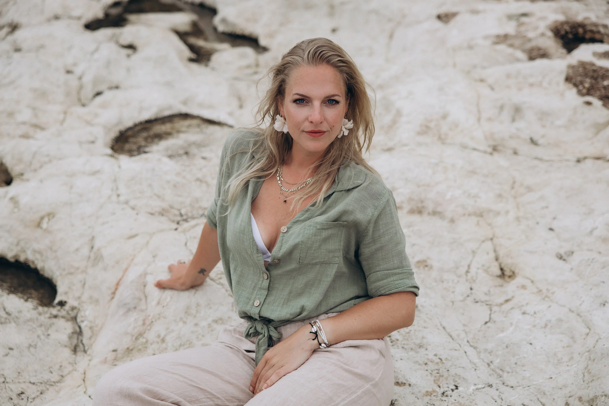 Een vrouw met blond haar zit op een rotsachtige ondergrond, draagt een lichtgroene blouse, witte broek en sieraden, en kijkt recht naar de camera met een rustige uitdrukking.