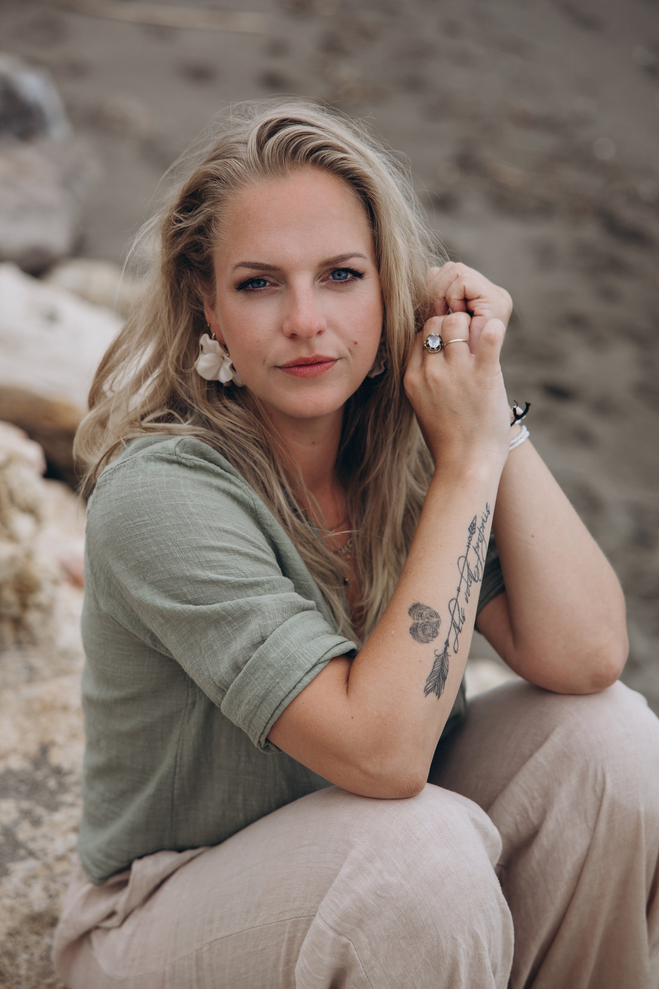 Een vrouw met lange blonde haren die op een rotsachtige ondergrond zit, haar rechter elleboog op haar knie, en haar rechter hand onder haar kin, in een natuurlijke omgeving.