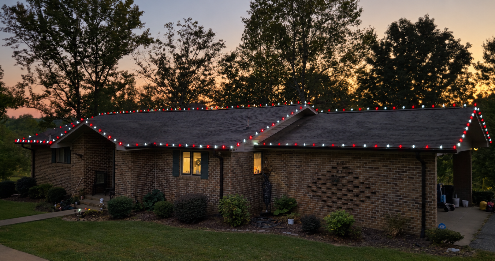 A brick house decorated with red and white Christmas lights on the roofline during sunset, with trees in the background.