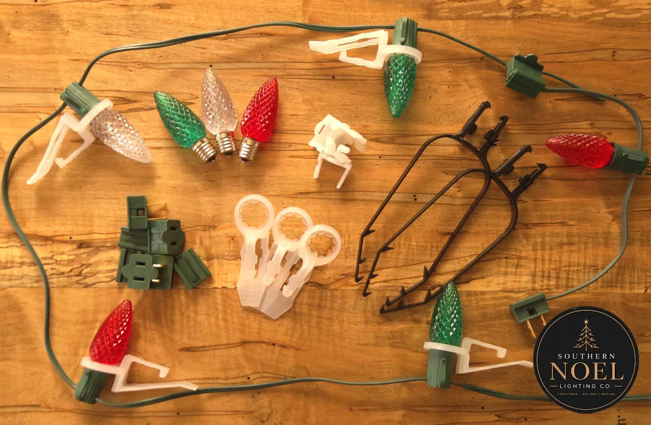 A string of Christmas holiday lights with colored bulbs, including red, green, and clear, lying on a wooden surface, with some plastic and metal clips and connectors scattered around.