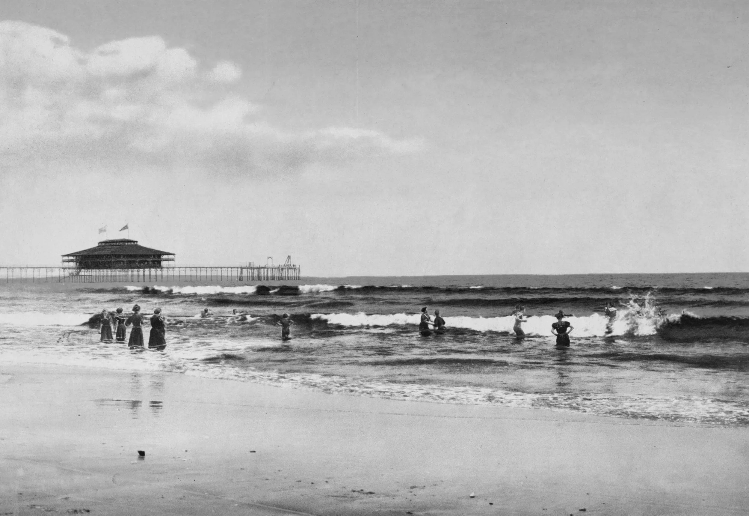 Black and white photo of people in the ocean near a pier with a building on stilts at the end of it, and cloudy sky.