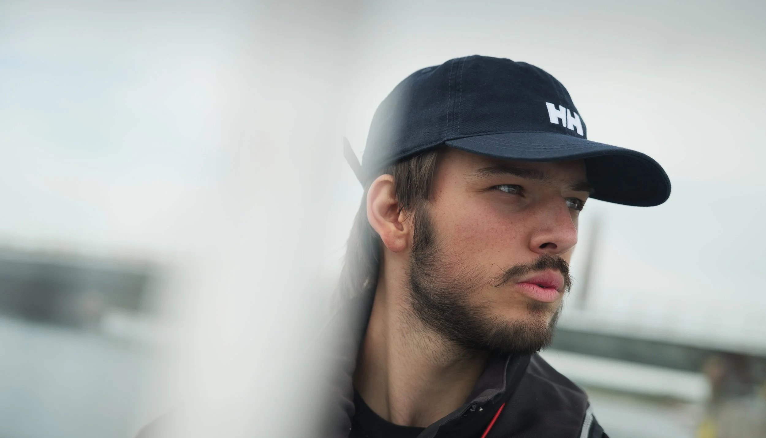 Close-up of a young man wearing a black cap with white text and a black jacket, looking to the side with a serious expression.
