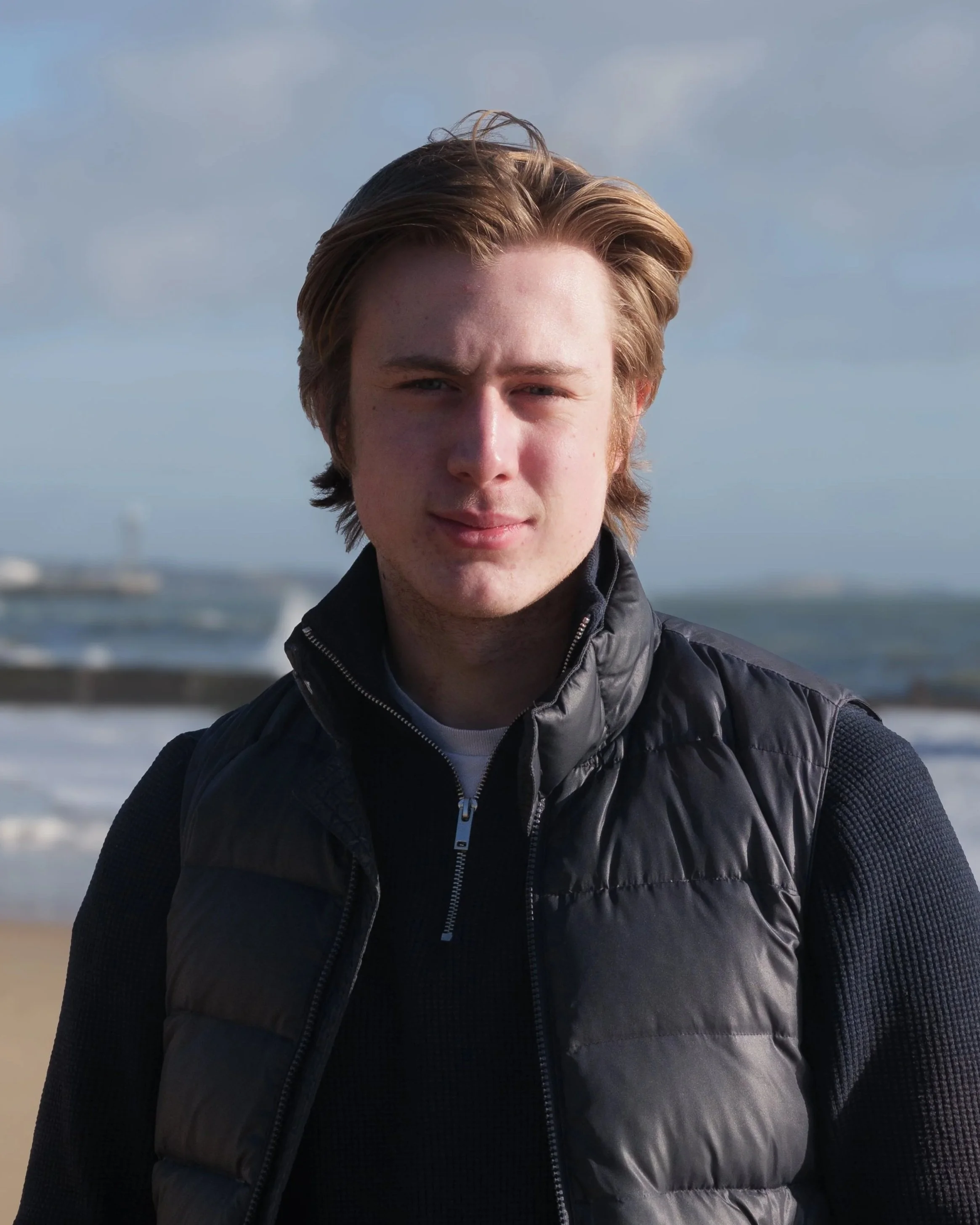 Close-up of a young man with blonde hair, wearing a black quilted jacket, standing outdoors near water on a cloudy day.