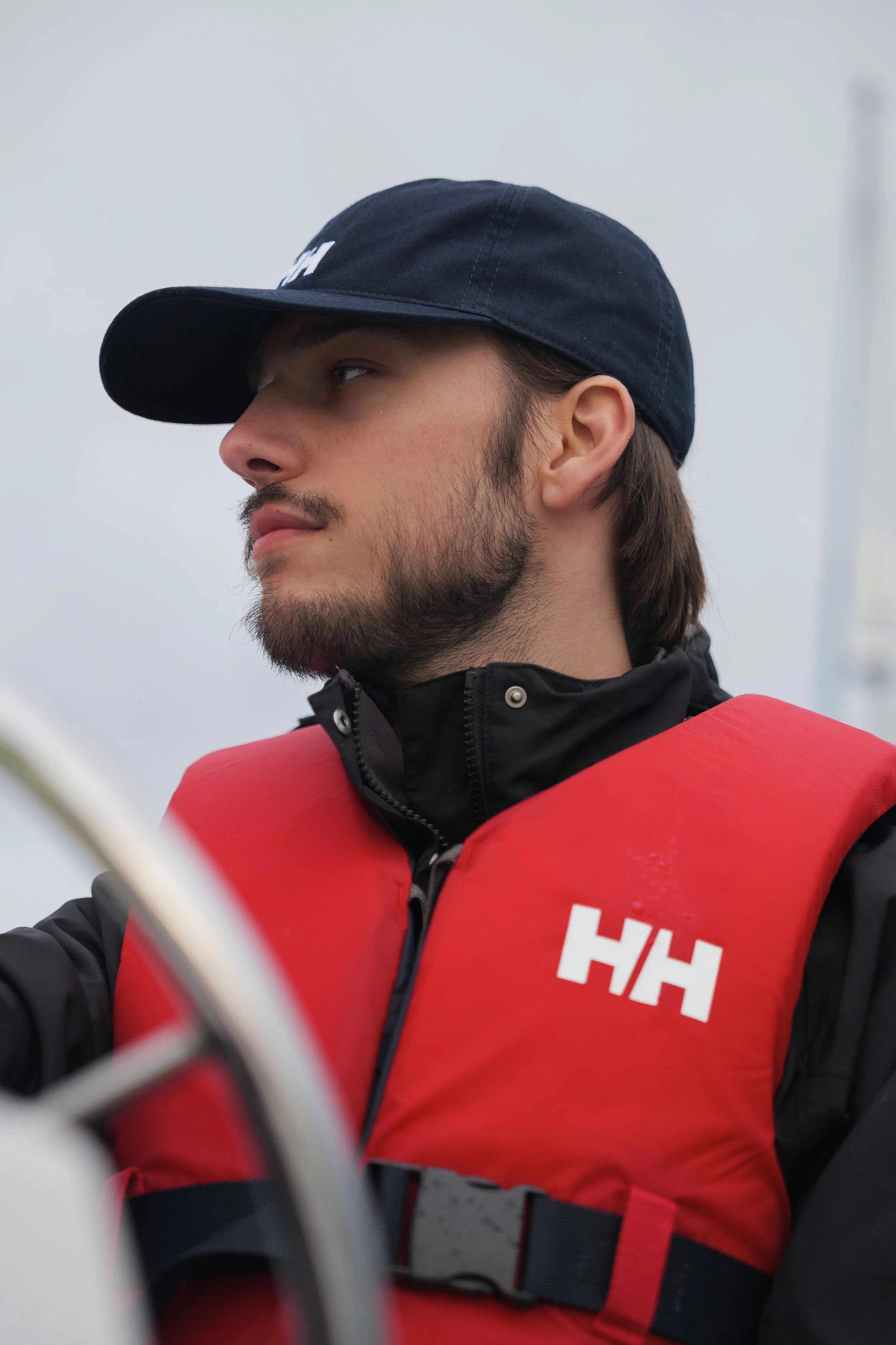 A young man wearing a black baseball cap and a red life jacket with the letters 'HH' on it. He appears to be on a boat or a similar watercraft, with a neutral, cloudy sky in the background.