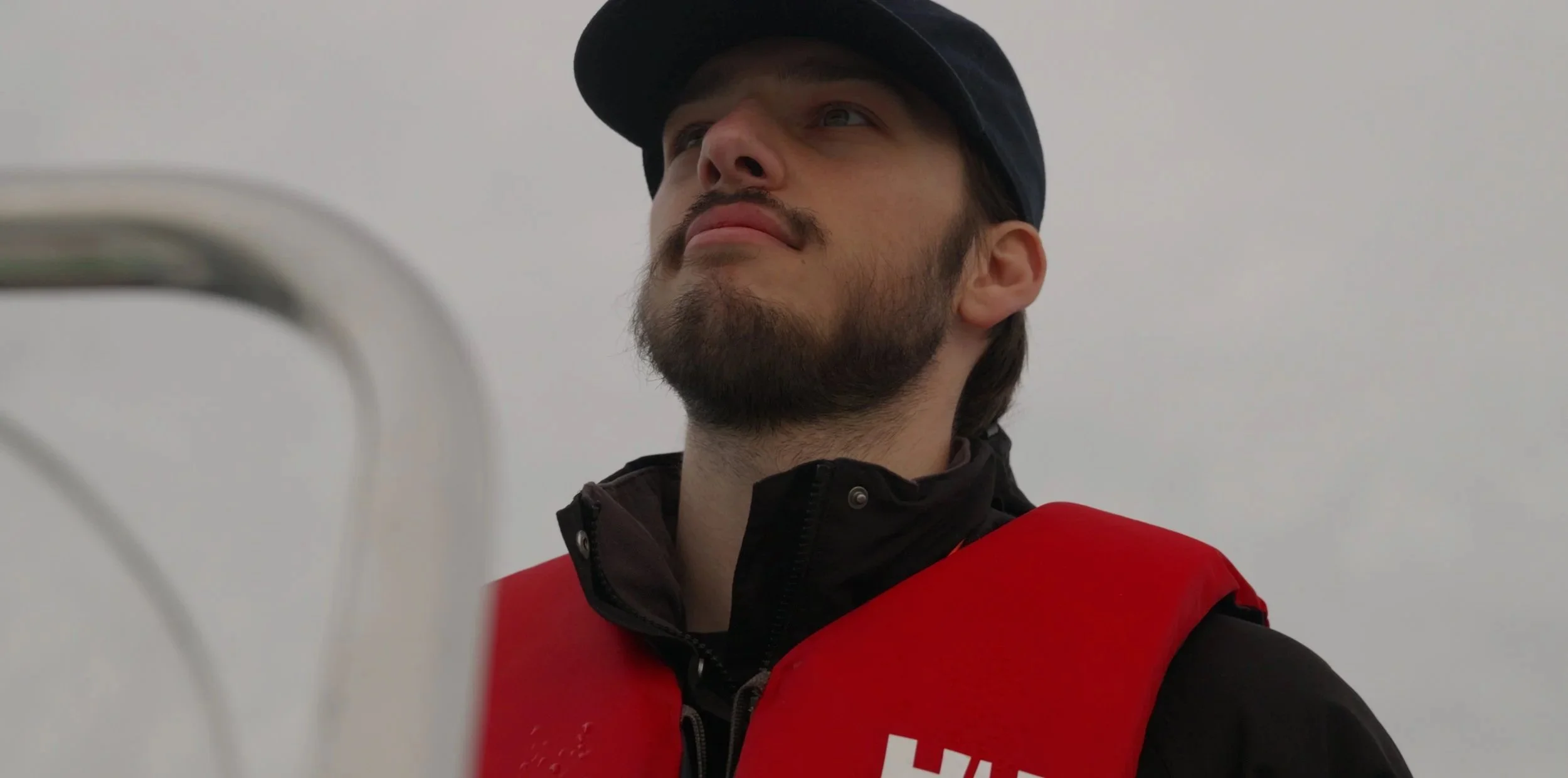 A man with a beard wearing a black cap and red vest looking upwards, with a white background.