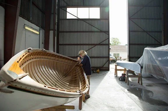 A person inspecting the interior of a wooden boat inside a large industrial workshop with an open door showing the outdoors.