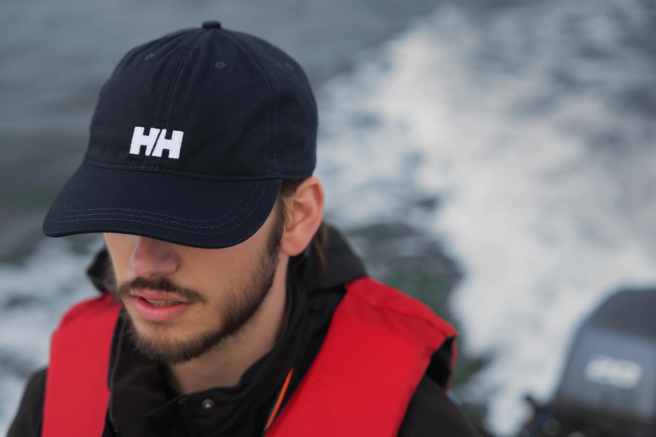 A man wearing a black baseball cap with white initials 'HH' and a red and black jacket, standing outdoors with a blurred ocean or water background.
