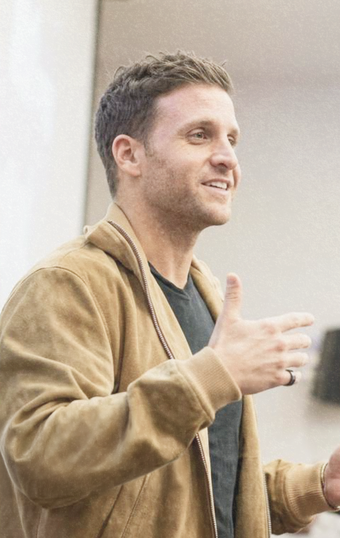 A man with short brown hair and a light beard, wearing a tan jacket over a black shirt, smiling and gesturing with his right hand.