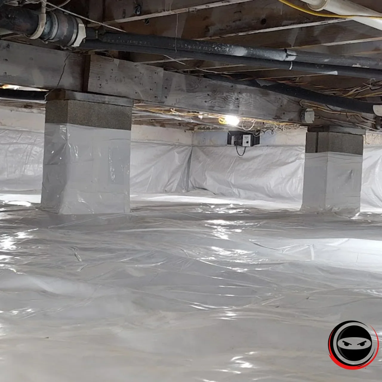Lower basement or crawl space with wooden beams, pipes, electrical wiring, and a plastic vapor barrier on the floor, supported by concrete blocks wrapped in plastic.