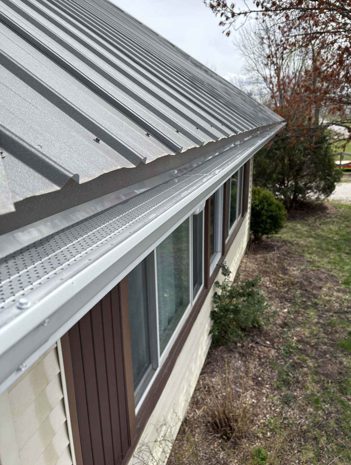 Close-up of a metal roof overhang on a house with trees and grass in the background.