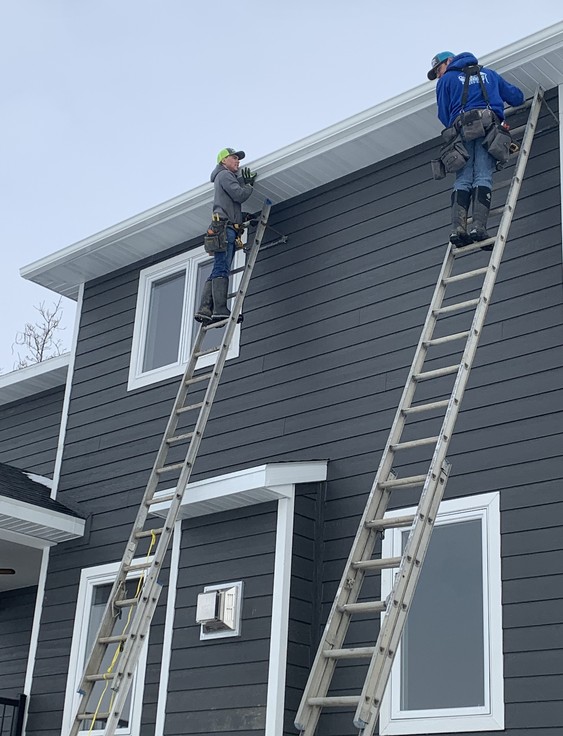 Two workers on ladders installing or repairing the eaves of a gray house with white trim and a window, wearing work gear and tools.