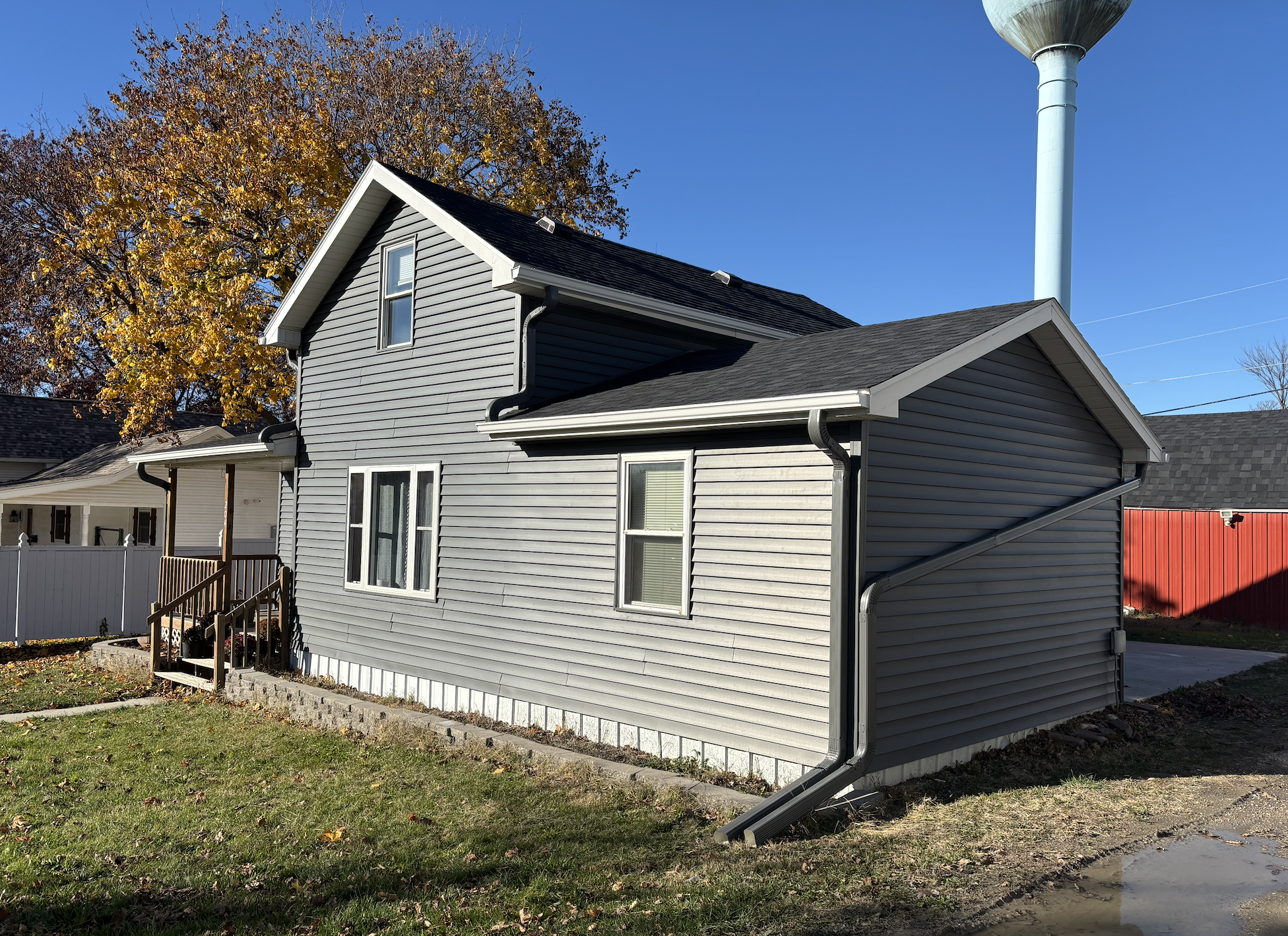 A gray house with white trim, a black roof, and a small porch with wooden steps. A large tree with yellow and brown leaves is in the background, along with a blue sky. There is a white fence, a red shed, and a water tower in the distance.
