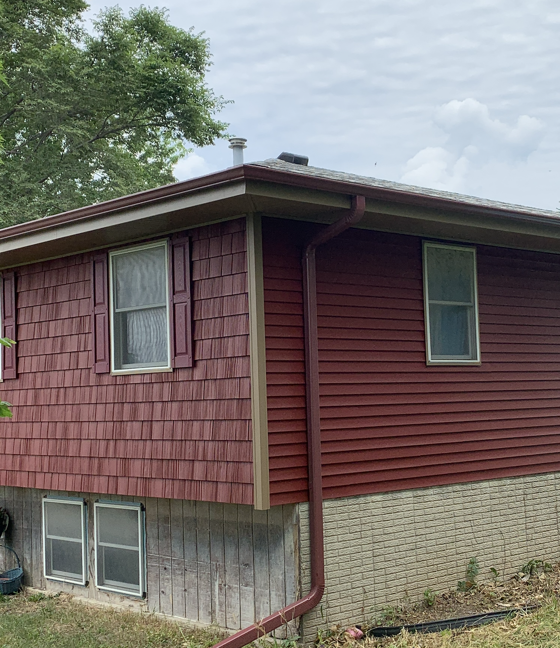 A two-story house with red horizontal siding and beige brick foundation. The upper floor has two windows with white frames and purple shutters, and the lower floor has two small windows near the ground. The roof has a vent and a chimney, and a red ra