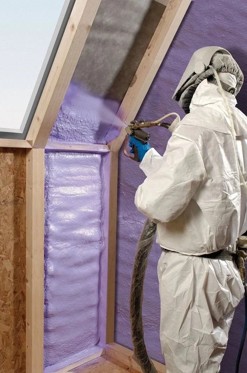 Person wearing protective gear spray foams onto a wall inside a framed section of a room under construction.