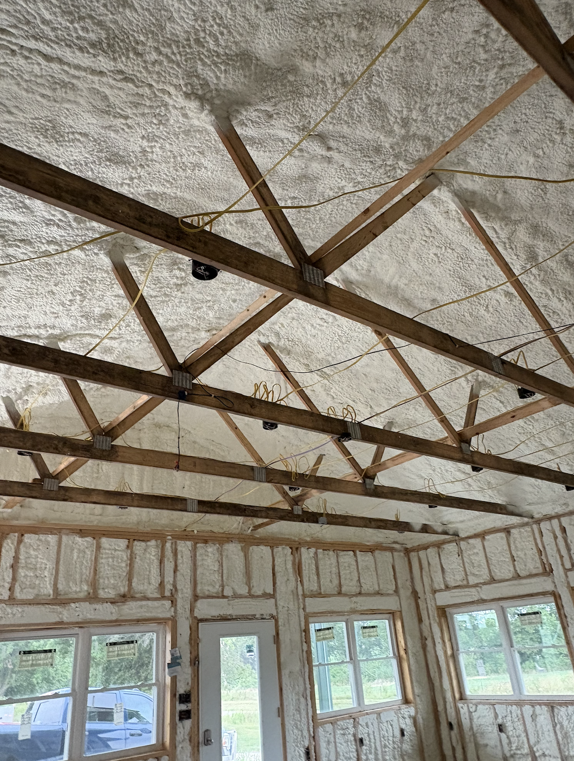 Interior room with exposed wooden ceiling joists, insulation foam sprayed on ceiling and walls, wiring hanging from ceiling, and multiple windows along the wall.