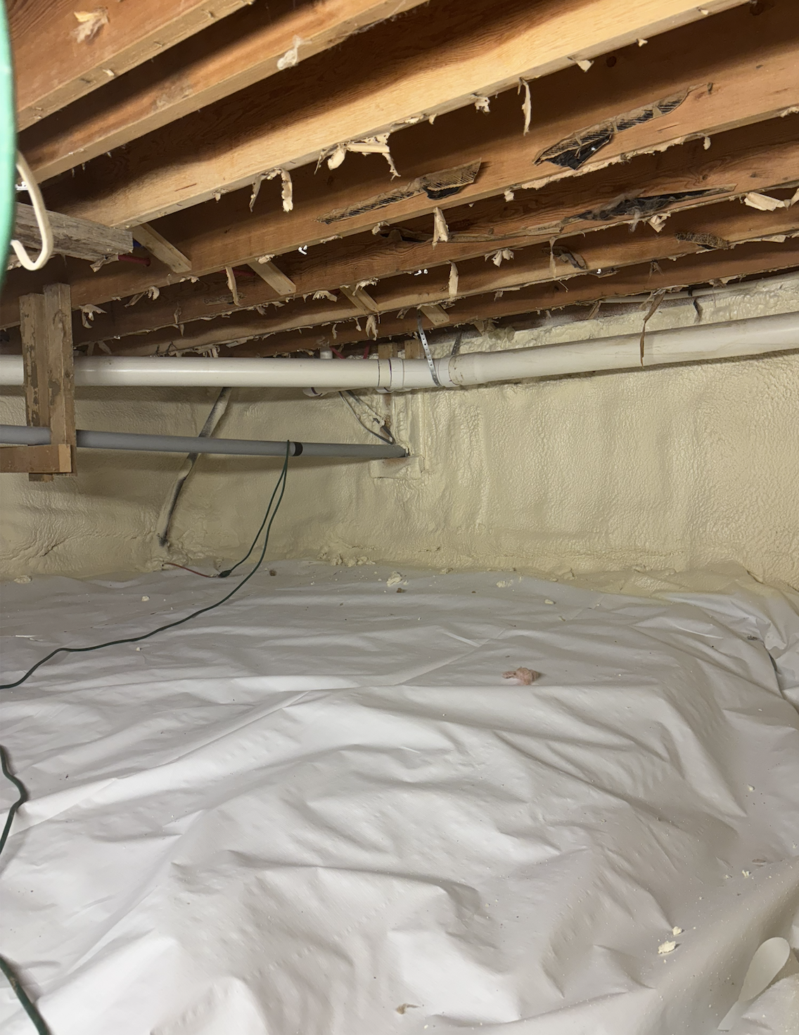 Underground area with foam spray insulation on the walls, exposed wooden ceiling beams, and PVC pipes, with plastic sheeting on the floor.