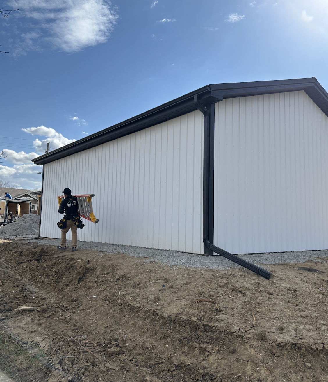 A construction worker standing on dirt ground holding a ladder against the side of a white building with black trim under a blue sky with clouds.