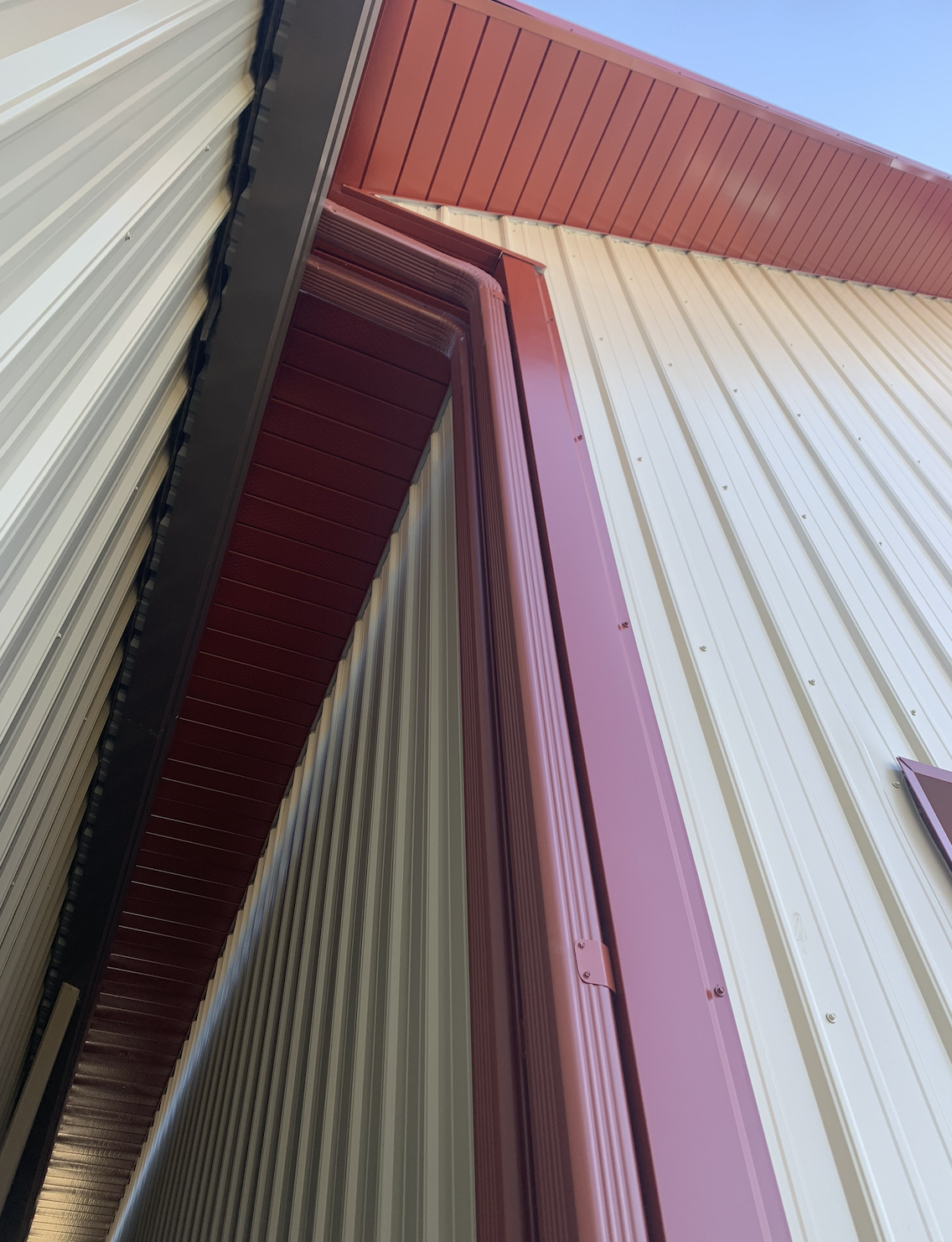 Close-up of the corner of a metal building with beige and maroon-colored siding, showing a maroon metal door frame and a maroon soffit.