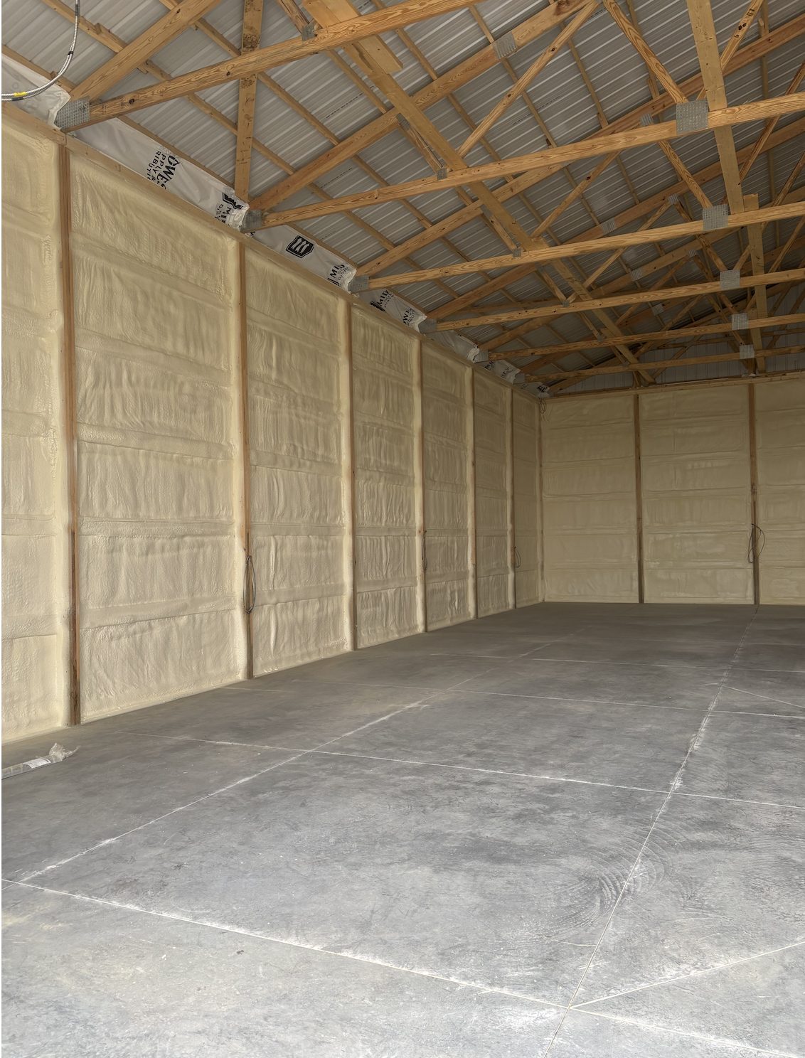 Empty garage or workshop with foam insulation on the walls, exposed wooden roof trusses, and a concrete floor.